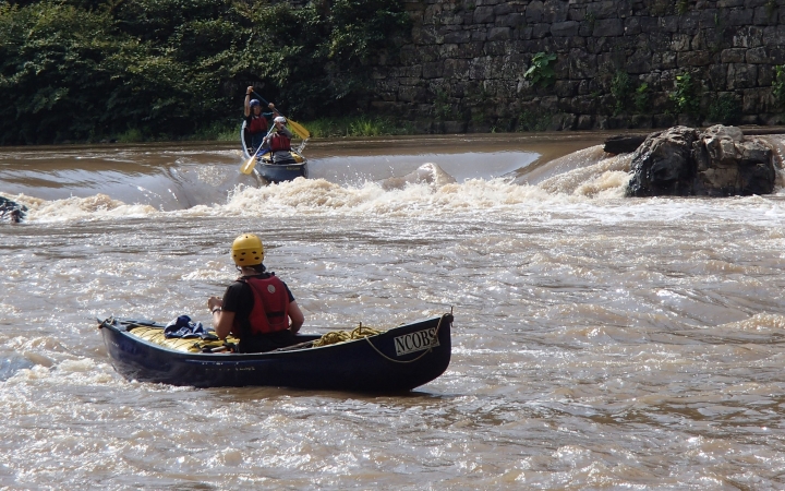 One person wearing safety gear sits in a canoe, watching two other people wearing safety gear paddle a canoe over a whitewater rapid.